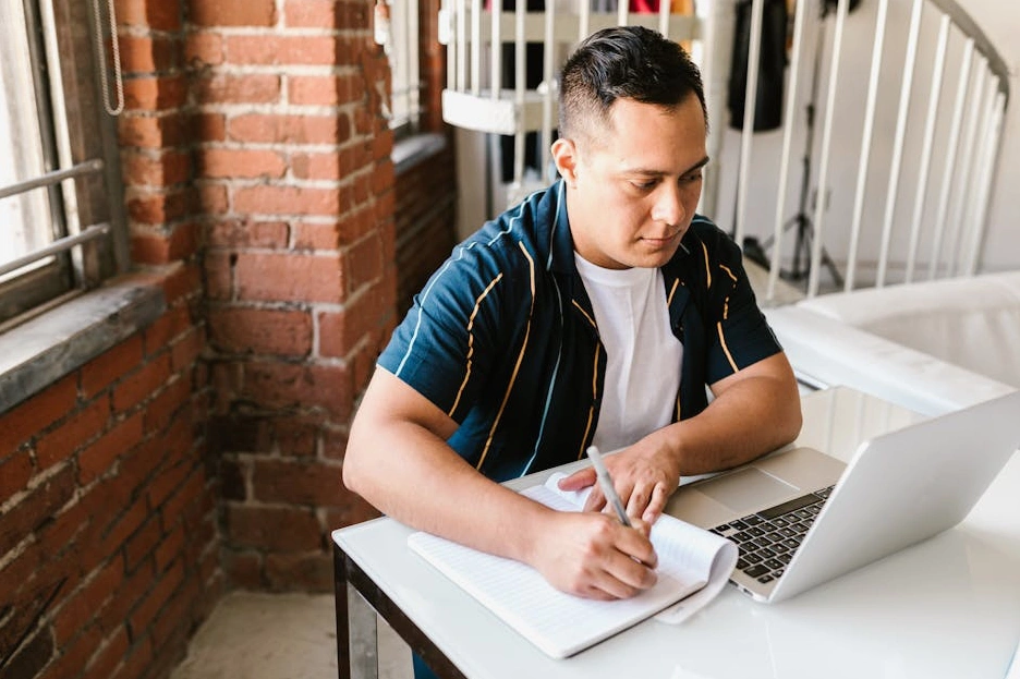 A person writing down financial goals and plans in a notebook with a laptop nearby.